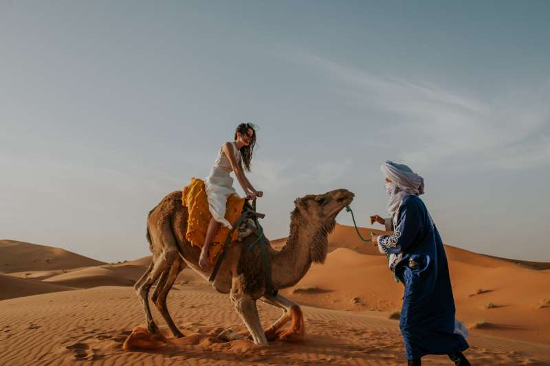 Girl on a camel trek in the Sahara desert of Morocco, with a native Berber helping her.
