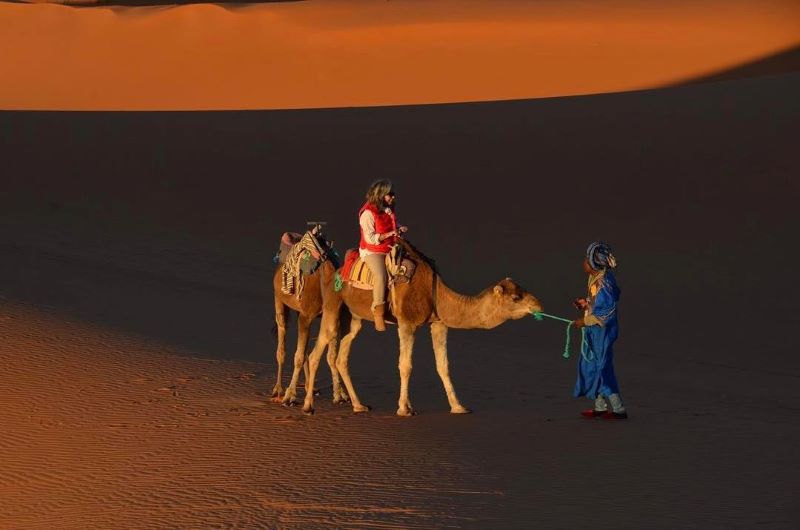 Woman riding a camel in the sunset in the desert in Morocco Woman riding a camel in the sunset while the guide is helping them both out, the camel and the woman